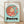 May include: Framed Studio Ghibli Ponyo poster print in light wood frame, leaning against white wall on wooden floor. Features circular illustration of red-haired girl Ponyo in coral dress floating underwater with goldfish, set against turquoise background. Bold black "PONYO" title below artwork with "A Studio Ghibli Film" text above. Minimalist room setting with soft natural lighting casting diagonal shadows on white textured wall.