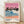 May include: Framed vintage-style beach poster showing pink sunset sky, dark silhouetted palm trees, blue ocean waves with white foam, coral-pink sand, and two small walking figures. Features color palette dots and Tokyo 1973 text below artwork. Light wooden frame displayed on wooden floor against white textured wall with soft shadows.