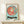May include: Framed Studio Ghibli Ponyo poster displayed on light wood shelf against cream wall. Poster features red-haired Ponyo character in circular frame on turquoise background with "PONYO" text below. Small wrapped white gift box with twine sits on shelf in front of natural wood frame.
