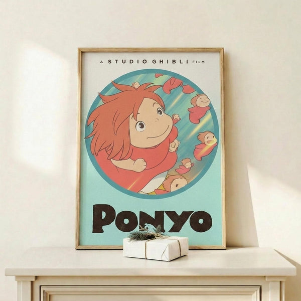 May include: Framed Studio Ghibli Ponyo poster displayed on light wood shelf against cream wall. Poster features red-haired Ponyo character in circular frame on turquoise background with "PONYO" text below. Small wrapped white gift box with twine sits on shelf in front of natural wood frame.