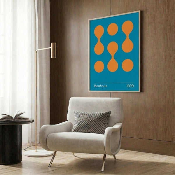 May include: Blue poster with orange geometric circles and "Bauhaus 1919" text in thin wooden frame, hanging on wood-paneled wall above cream upholstered armchair with geometric patterned pillow, brass floor lamp and round black side table with open book nearby