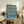 May include: Framed blue poster with orange geometric circle and oval shapes arranged in a 3x3 grid, displaying "Bauhaus 1919" text at bottom. Shows natural wood frame leaning against white wall on rustic wooden sideboard alongside fern plant, ceramic vases, and stacked books in cozy living room setting with leather armchair visible.