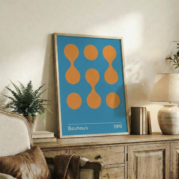 May include: Framed blue poster with orange geometric circle and oval shapes arranged in a 3x3 grid, displaying "Bauhaus 1919" text at bottom. Shows natural wood frame leaning against white wall on rustic wooden sideboard alongside fern plant, ceramic vases, and stacked books in cozy living room setting with leather armchair visible.