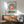 May include: Framed Ponyo poster displayed above a wooden sideboard in a mid-century modern living room. The artwork features the red-haired character in a circular teal design with "PONYO" in bold black letters below. Natural sunlight creates dramatic shadows on white walls. A teal upholstered armchair with wooden frame sits beside a round coffee table, with a small white table lamp on the credenza.