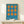 May include: Framed Bauhaus-style poster with blue background and orange geometric pattern of circles and connected oval shapes, displayed on light wood surface next to small wrapped gift box with twine and greenery accent against white wall