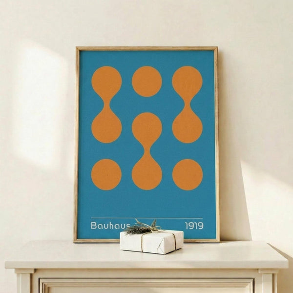 May include: Framed Bauhaus-style poster with blue background and orange geometric pattern of circles and connected oval shapes, displayed on light wood surface next to small wrapped gift box with twine and greenery accent against white wall