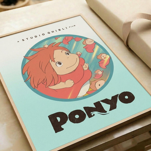 Light blue poster print featuring Ponyo, the red-haired girl character from Studio Ghibli, inside a circular frame surrounded by small floating figures. The character has orange-red hair and wears a red outfit. "PONYO" appears in large black letters at the bottom, with "STUDIO GHIBLI FILM" text at the top. The print sits on a cream-colored surface next to rolled white paper in soft, warm lighting.