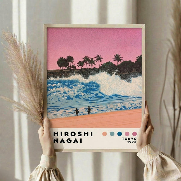May include: Person holding framed poster print featuring tropical beach scene with pink sunset sky, palm trees, blue ocean waves, and two small figures on sandy shore. Artwork displays retro color palette with coral, teal, and pink tones. Light wooden frame against neutral curtained background with dried pampas grass visible to left side.