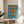 May include: A framed Bauhaus-inspired poster with bright blue background and nine orange geometric shapes arranged in a 3x3 grid, featuring connected circles and oval forms. The minimalist print sits on a light wood round side table next to a beige upholstered chair in a sunlit room with white walls and a plant.