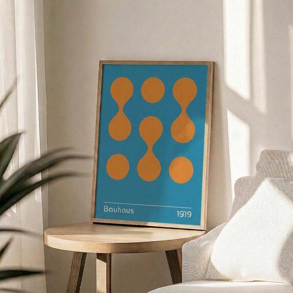 May include: A framed Bauhaus-inspired poster with bright blue background and nine orange geometric shapes arranged in a 3x3 grid, featuring connected circles and oval forms. The minimalist print sits on a light wood round side table next to a beige upholstered chair in a sunlit room with white walls and a plant.