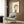 May include: Framed poster print featuring a stylized cartoon child with brown hair wearing an orange shirt, displayed on wood paneled wall above a beige upholstered armchair with geometric patterned pillow. Modern reading corner with brass floor lamp, dark side table with open book, and natural lighting from window with curtains.