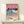 May include: Framed poster print displayed on light wood shelf against cream wall. Artwork shows tropical beach scene with blue ocean waves, coral pink sand, silhouetted palm trees against pink sunset sky, and two small figures on shore. Text reads "HIROSHI NAGAI" and "TOKYO 1973" with colored dots below. Small wrapped gift box with evergreen sprig sits beside thin gold frame.