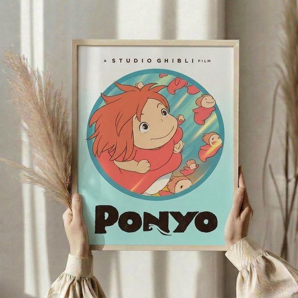 May include: Person holding framed Studio Ghibli Ponyo poster in light wood frame against white curtained window. Poster features circular illustration of red-haired Ponyo character surrounded by small fish on turquoise background with black "PONYO" text below. Person wears cream ruffled sleeves, holds dried pampas grass stems in left hand.