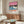 May include: Framed poster showing tropical beach scene with pink sky, palm trees, blue waves, coral sand, and two silhouetted figures. Displayed above wooden mid-century credenza with white table lamp. Room features cream walls, white upholstered swivel chair, oval glass coffee table, and natural lighting creating warm shadows across polished floor.