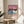 May include: Framed poster artwork displaying a tropical beach scene with pink sunset sky, palm tree silhouettes, blue ocean waves, coral-colored sand, and two small figures walking on the beach. The print features "HIROSHI NAGAI" and "TOKYO 1973" text with colorful dots. Shown in thin wooden frame on round wooden side table next to beige upholstered chair with white throw pillow, green plant leaves visible, soft natural lighting creates shadows on white wall.