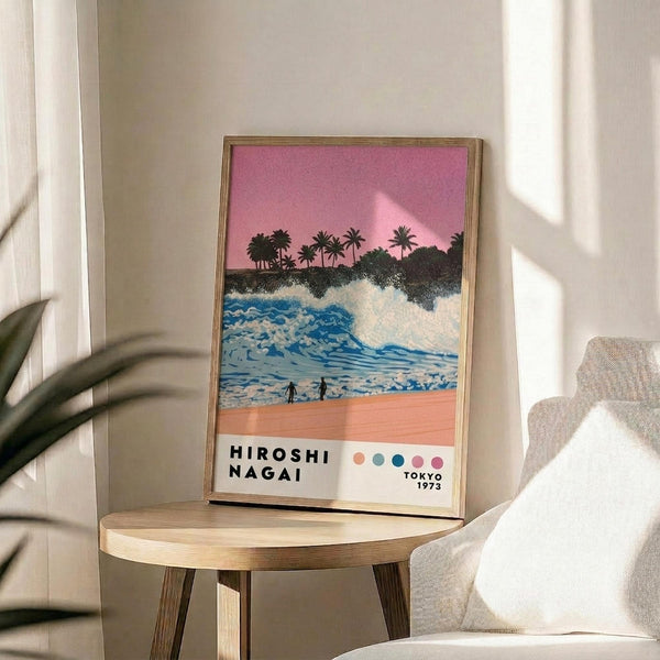 May include: Framed poster artwork displaying a tropical beach scene with pink sunset sky, palm tree silhouettes, blue ocean waves, coral-colored sand, and two small figures walking on the beach. The print features "HIROSHI NAGAI" and "TOKYO 1973" text with colorful dots. Shown in thin wooden frame on round wooden side table next to beige upholstered chair with white throw pillow, green plant leaves visible, soft natural lighting creates shadows on white wall.