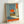 May include: Framed poster leaning against white wall showing surreal scene viewed through open doorway. Interior has tiled floor and blue-gray walls. Through doorway, silhouetted figure sits on grassy hill overlooking water with large orange sun in cloudy sky. Orange text box with Japanese characters in upper right corner. Orange text band across bottom. Light wood frame with natural lighting creating soft shadows.