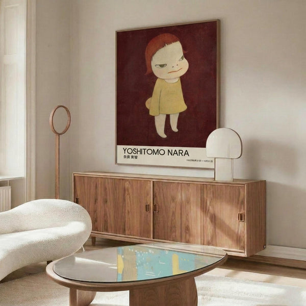 May include: Yoshitomo Nara exhibition poster featuring a pale child with reddish hair in yellow dress on dark red background, displayed above a walnut wood sideboard in modern living room. White curved chair and glass-top coffee table with colorful art visible. Minimalist interior with cream walls and natural lighting.