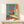 May include: Framed poster displaying a surreal scene of a person sitting on golden ground viewing a large orange sun through an open doorway, with Japanese text on orange banner, styled in vintage travel poster aesthetic. Small white wrapped gift sits beside the wooden frame on light wood surface against white wall.