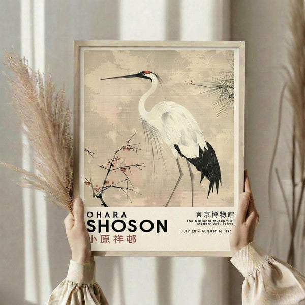 May include: Person holding a framed vintage Japanese art print featuring a white crane with black wing tips and long curved neck, standing among delicate pink flowering branches and pine needles on a beige textured background. Exhibition poster text appears below the artwork. Natural lighting illuminates the cream-colored wall and dried pampas grass in a minimalist interior setting.
