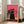 May include: Framed poster with pink background showing black cat wearing yellow sunglasses and collar in center, surrounded by colorful cats in teal sunglasses - some black, pink, and turquoise. Cats sit on yellow and orange wooden surface. White frame leans against beige wall next to gray sofa in bright living room with windows.