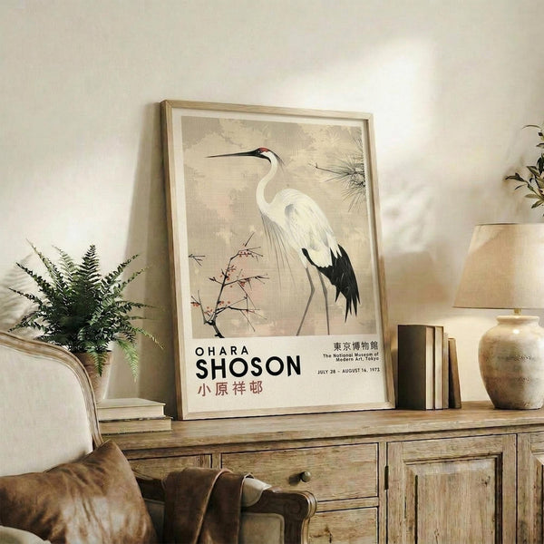 May include: Framed vintage Japanese crane artwork displayed on rustic wooden sideboard in neutral-toned living room. Print features elegant white heron with black wing tips and red head marking against beige background with botanical elements. Exhibition poster format with text below bird illustration. Scene includes fern plant, ceramic vase, stacked books, and cream upholstered furniture.