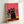 May include: Framed poster print featuring stylized cats wearing sunglasses against a coral pink background. The main black cat in center wears yellow sunglasses and a collar, surrounded by pink, green, and black cats all sporting teal sunglasses. The cats sit on wooden planks with a yellow textured surface behind. The artwork is displayed in a light wood frame leaning against a white wall.