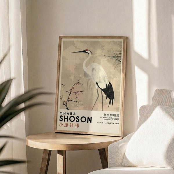 May include: Vintage Japanese crane art poster in thin wooden frame displayed on round wooden side table next to beige upholstered chair. Soft natural lighting creates gentle shadows on neutral wall. Green plant leaves visible in corner. Poster features elegant white crane with black wing tips and red head marking among delicate branches, with exhibition text below in muted earth tones.