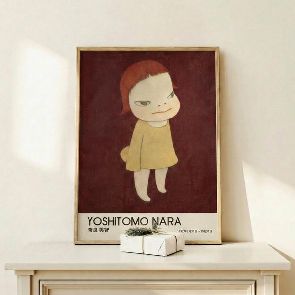 May include: A framed art poster featuring a pale child-like figure with reddish hair wearing a yellow dress against a deep burgundy background, displayed on a light wood shelf next to a small wrapped gift with evergreen sprig. The poster shows Japanese text and exhibition dates from 1992. Minimalist styling with white walls and natural lighting.