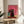 May include: Framed poster print featuring a black cat wearing sunglasses and collar standing prominently in front of several other colorful cats, all wearing sunglasses, against a vibrant pink background. The illustration shows cats in various colors including teal, pink, and black in a stylized, cartoon-like art style. The artwork is displayed in a thin wooden frame on a round wooden side table next to a beige upholstered chair, with natural sunlight streaming across white walls and a plant vi