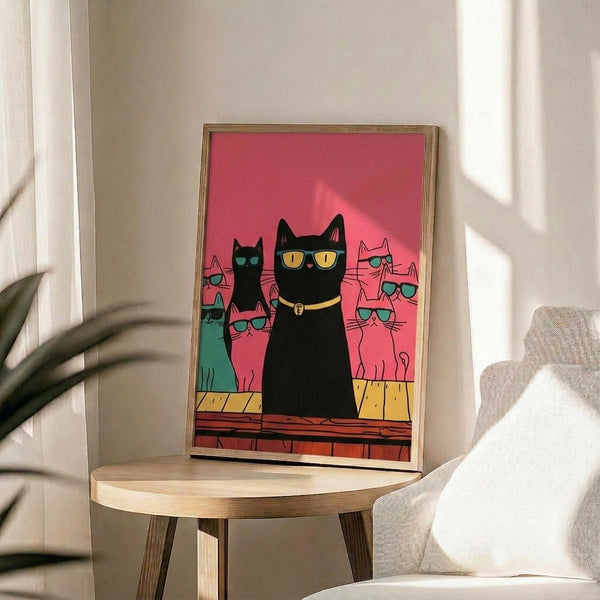 May include: Framed poster print featuring a black cat wearing sunglasses and collar standing prominently in front of several other colorful cats, all wearing sunglasses, against a vibrant pink background. The illustration shows cats in various colors including teal, pink, and black in a stylized, cartoon-like art style. The artwork is displayed in a thin wooden frame on a round wooden side table next to a beige upholstered chair, with natural sunlight streaming across white walls and a plant vi