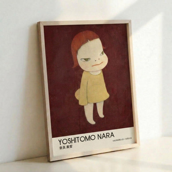 May include: Framed poster leaning against white wall showing stylized illustration of pale child with reddish-brown hair wearing yellow dress on deep burgundy background. White border at bottom displays "YOSHITOMO NARA" text with Japanese characters and dates. Light wood frame, soft natural lighting creates gentle shadows on wall.