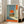May include: Framed poster leaning against beige couch in bright living room with large windows. Artwork shows person sitting in doorway overlooking landscape with large orange sun, rendered in retro illustration style with muted blue and orange tones. Japanese text appears on orange vertical banner. White matted frame sits on light wood floor next to neutral furniture.