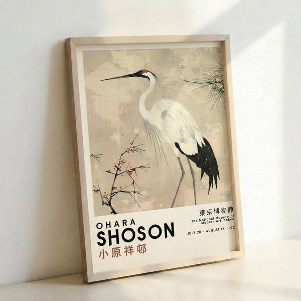 May include: Framed museum exhibition poster featuring a white crane with black wing tips and long beak standing among delicate flowering branches and pine needles on a muted beige background. The artwork shows Japanese-style nature illustration with text reading "OHARA SHOSON" and Japanese characters below, plus museum details. Light wood frame leaning against white wall on light surface.