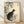 May include: Framed vintage Japanese woodblock print featuring a black and white cat sitting beneath flowering branches with green leaves and pale blossoms. The artwork has a cream background with red signature stamps, displayed in a light wood frame against white walls on wooden flooring with natural window light creating soft shadows.