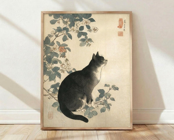 May include: Framed vintage Japanese woodblock print featuring a black and white cat sitting beneath flowering branches with green leaves and pale blossoms. The artwork has a cream background with red signature stamps, displayed in a light wood frame against white walls on wooden flooring with natural window light creating soft shadows.