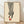 May include: Framed fashion illustration print leaning against white textured wall on light wood floor. Cream-colored poster shows black and white striped wide-leg pants with one foot wearing a red high heel sandal. Simple line art style in thin natural wood frame with subtle shadows cast on wall.