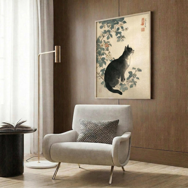 May include: Japanese-style cat artwork print in light wooden frame hanging on wood-paneled wall above beige upholstered armchair with geometric patterned pillow. Print shows black and white cat sitting among flowering vine branches in traditional Asian art style with muted green, cream and coral tones. Modern brass floor lamp and dark round side table with open book complete the neutral-toned reading nook scene.