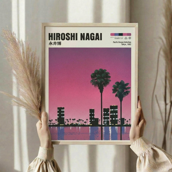 May include: Hands holding a framed poster print in a light wood frame, featuring a stylized cityscape silhouette with palm trees against a vibrant pink and purple gradient sky, reflected in water below. The artwork shows a retro aesthetic with geometric buildings and tropical elements. Background shows dried pampas grass and neutral curtains in soft lighting.
