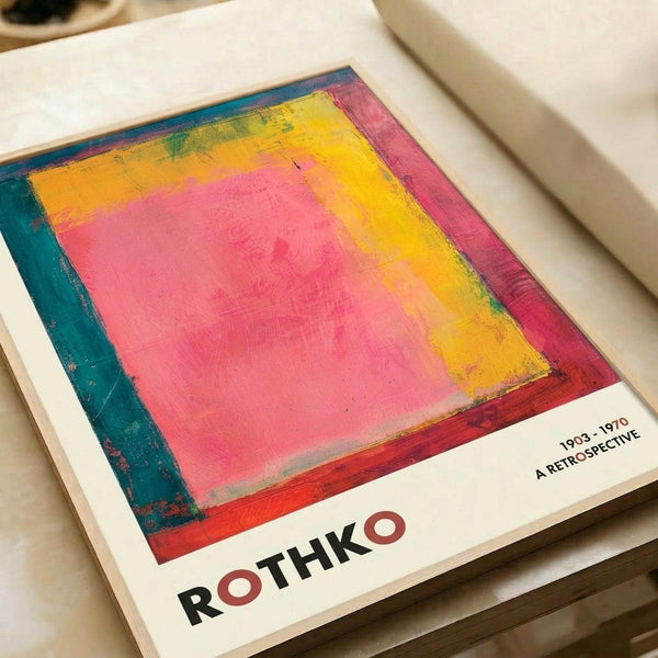Colorful abstract art book or catalog cover featuring a vibrant rectangular composition with pink center, yellow and orange surrounding areas, and teal border. The cover displays "ROTHKO" in bold black letters at bottom with red circular accents, and "1903-1970 A RETROSPECTIVE" text in upper right corner. Book lies open on white surface showing textured, painterly brushwork in warm and cool tones.
