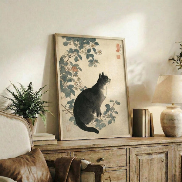 May include: Japanese woodblock print artwork featuring a gray and white cat sitting beneath flowering rose branches with green leaves, displayed in a light wood frame on a rustic wooden sideboard alongside fern plant, ceramic vase, and books in a neutral-toned living room setting