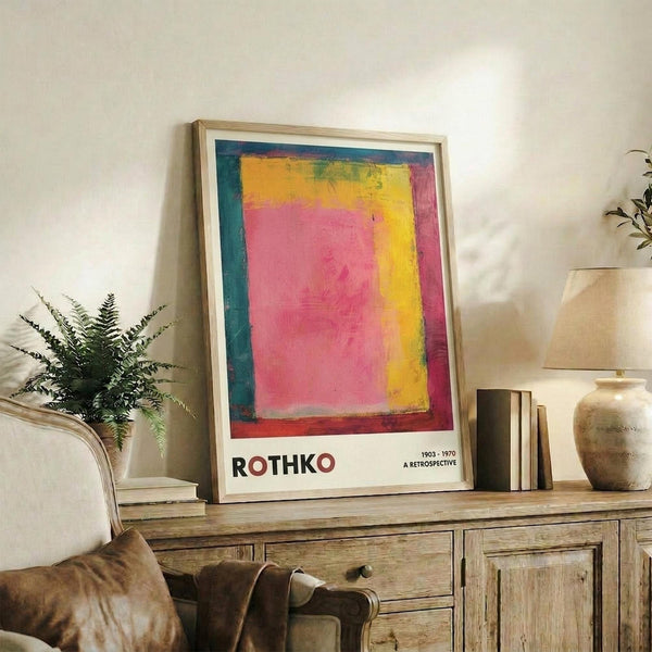 May include: Framed colorful abstract art poster displayed on rustic wooden sideboard in cozy living room setting. Artwork features vibrant pink, yellow, and teal color blocks with exhibition text below. Scene includes cream upholstered chair, potted fern plant, stacked books, ceramic vase, and neutral wall background with soft natural lighting.