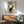 May include: Japanese woodblock print artwork featuring a black cat sitting among flowering rose branches, displayed in wooden frame above mid-century modern walnut credenza with ceramic table lamp, next to sage green upholstered armchair with wooden arms in bright sunlit room with white walls
