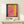 May include: Framed art print featuring abstract color blocks in pink, yellow, orange, teal and burgundy with white exhibition text below reading "ROTHKO 1903-1970 A RETROSPECTIVE." Light wood frame sits on white floating shelf against cream wall with small wrapped gift beside it.