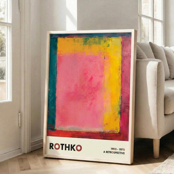 May include: A framed exhibition poster featuring an abstract painting with vibrant pink, yellow, orange and teal color blocks in Rothko's signature style. The white poster displays "ROTHKO" and "1903-1970 A RETROSPECTIVE" in dark text below the artwork. The thin wooden frame leans against a white wall next to a beige upholstered sofa in a bright, minimalist living room with large windows and natural light.