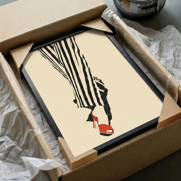 May include: Framed fashion illustration print in black frame, packed in brown cardboard box with tissue paper wrapping. Artwork shows stylized legs in black and white striped pants wearing red high heel shoes on cream background. Print appears medium-sized, positioned on white surface with wooden rolling pin nearby.