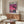 May include: Framed poster showing pink-purple sunset cityscape with palm trees and buildings reflected in water, displayed above mid-century modern walnut credenza in minimalist living room with white curved chair, brass floor lamp, white table lamp, and glass-top coffee table on light wood floors.