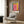 May include: Colorful abstract art poster with vibrant pink, yellow, blue, and red rectangular blocks in white frame, displayed above cream upholstered armchair with geometric patterned pillow. Modern interior setting features warm wood paneling, brass floor lamp, and dark round side table with open book.