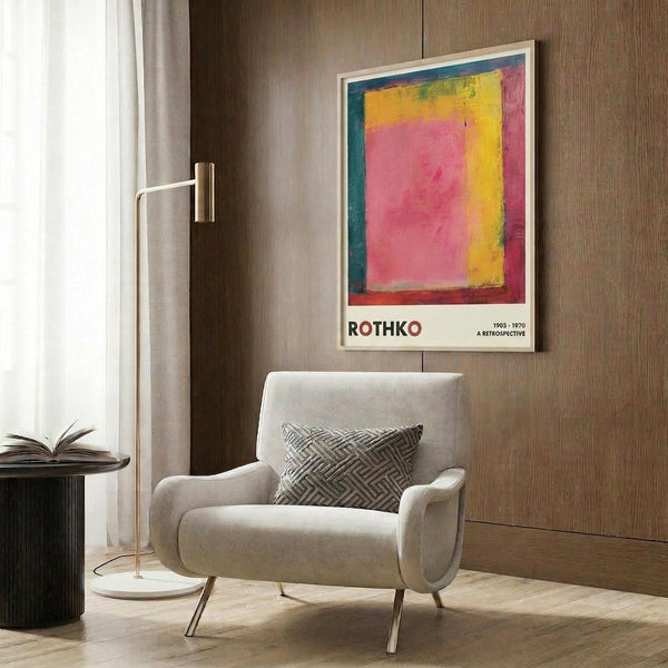 May include: Colorful abstract art poster with vibrant pink, yellow, blue, and red rectangular blocks in white frame, displayed above cream upholstered armchair with geometric patterned pillow. Modern interior setting features warm wood paneling, brass floor lamp, and dark round side table with open book.