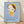 May include: Framed poster print leaning against white wall on light wooden floor. Shows stylized illustration of pale child figure with green eyes, brown bob haircut, yellow beret and dress against blue background. Text reads "Yoshitomo Nara" with Japanese characters and exhibition dates. Natural wood frame, soft lighting creates shadows on textured white wall.